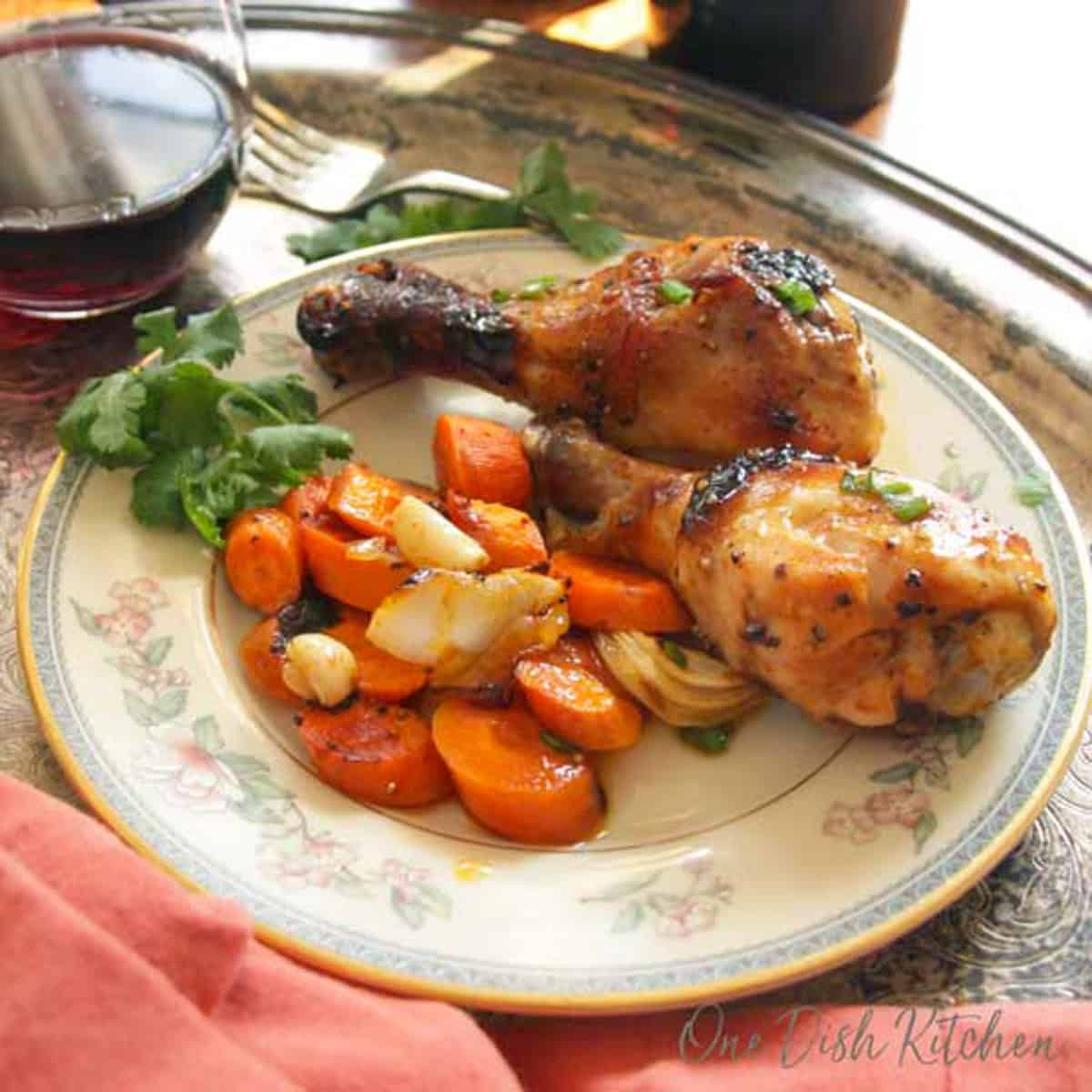 two baked chicken legs on a plate with vegetables and a glass of red wine.