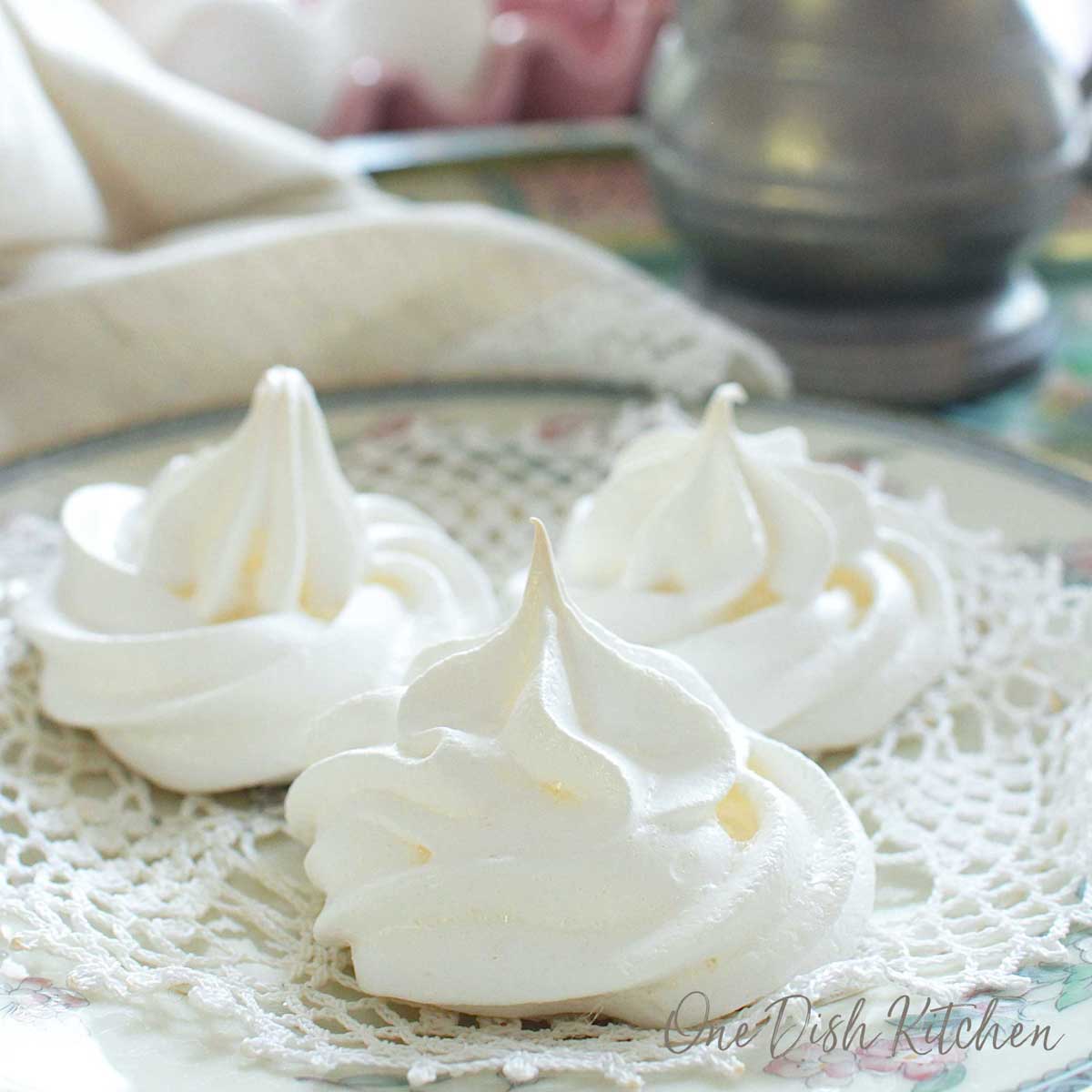 three meringue cookies on a doily on top of a white plate next to a beige napkin and eggs.