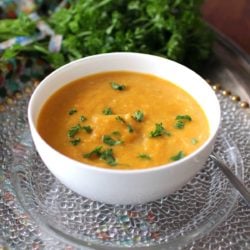 a white bowl filled with curried butternut squash soup on top of a clear plate next to parsley