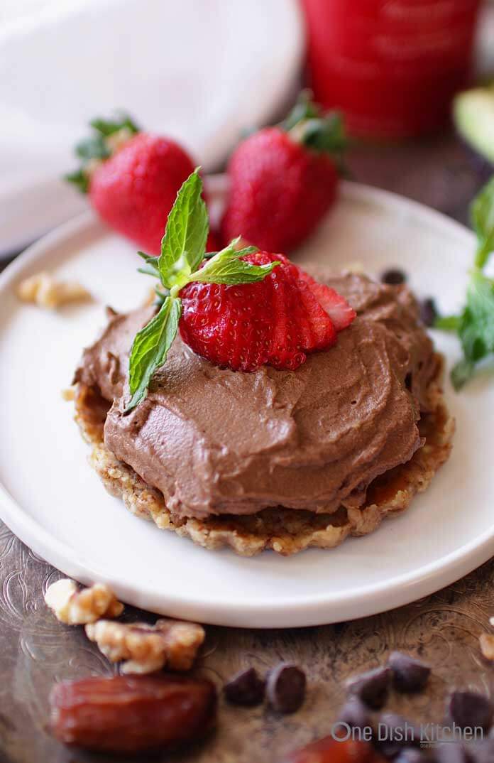 A single-serving Chocolate Avocado pie topped with a strawberry on a small plate surrounded by chocolate chips, walnut halves, dates, and strawberries 