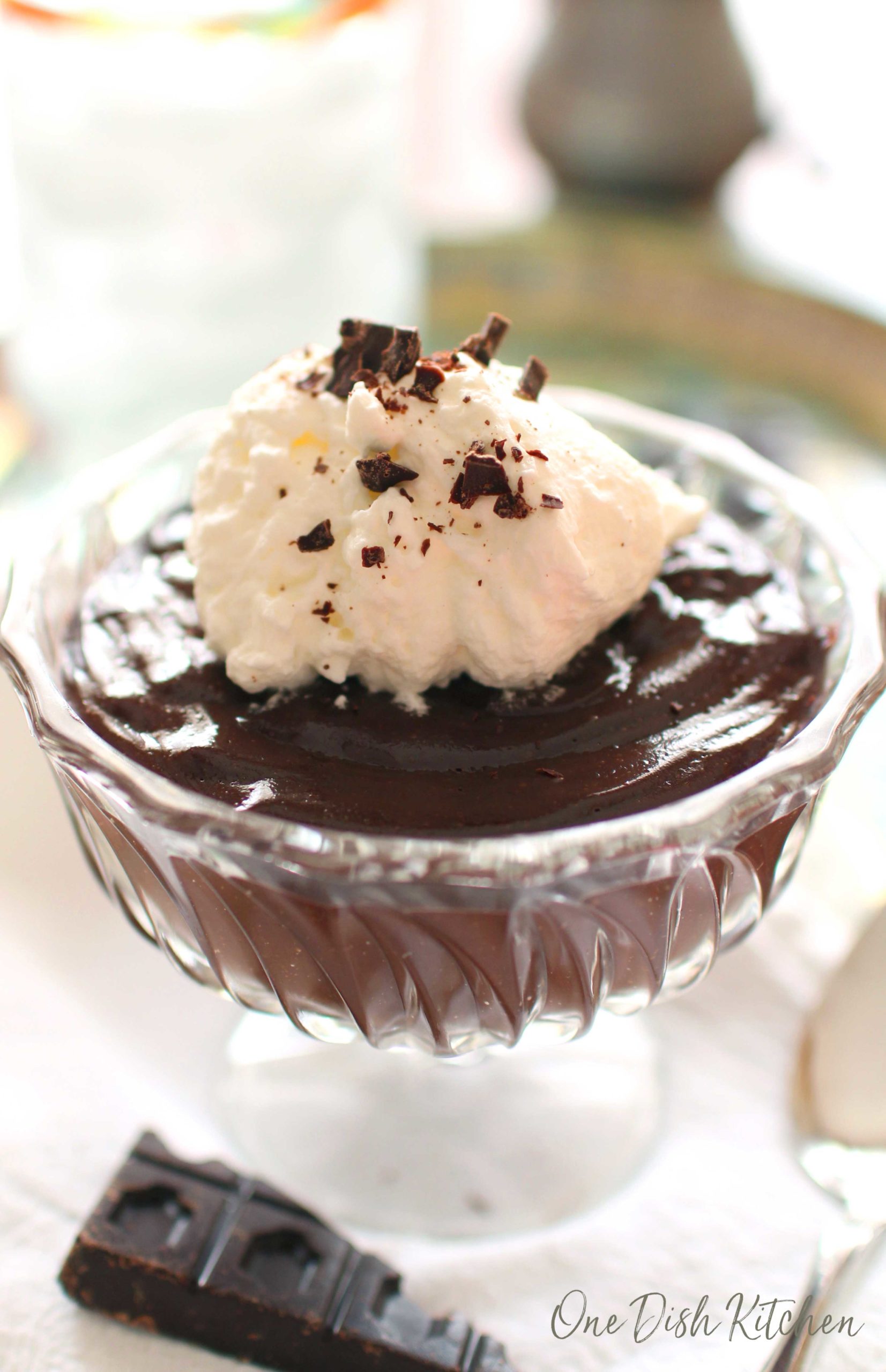 Chocolate pudding topped with whipped cream and chocolate shavings in a dessert glass next to a piece of a chocolate bar and a spoon.