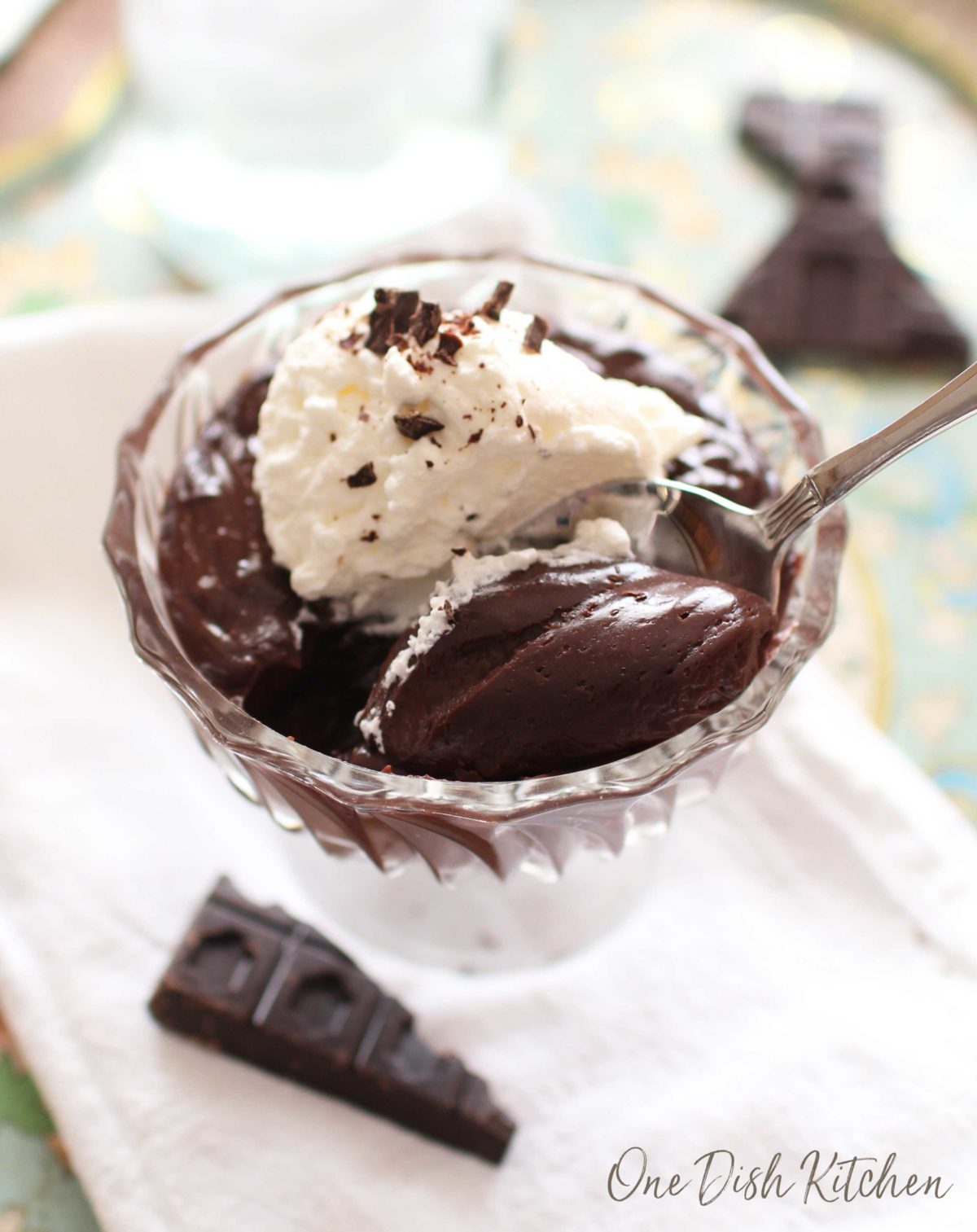 Overhead view of chocolate pudding topped with whipped cream and chocolate shavings in a dessert glass next to a piece of a chocolate bar and a spoon.