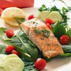 a piece of roasted salmon on a bed of spinach leaves surrounded by cherry tomatoes and cucumbers
