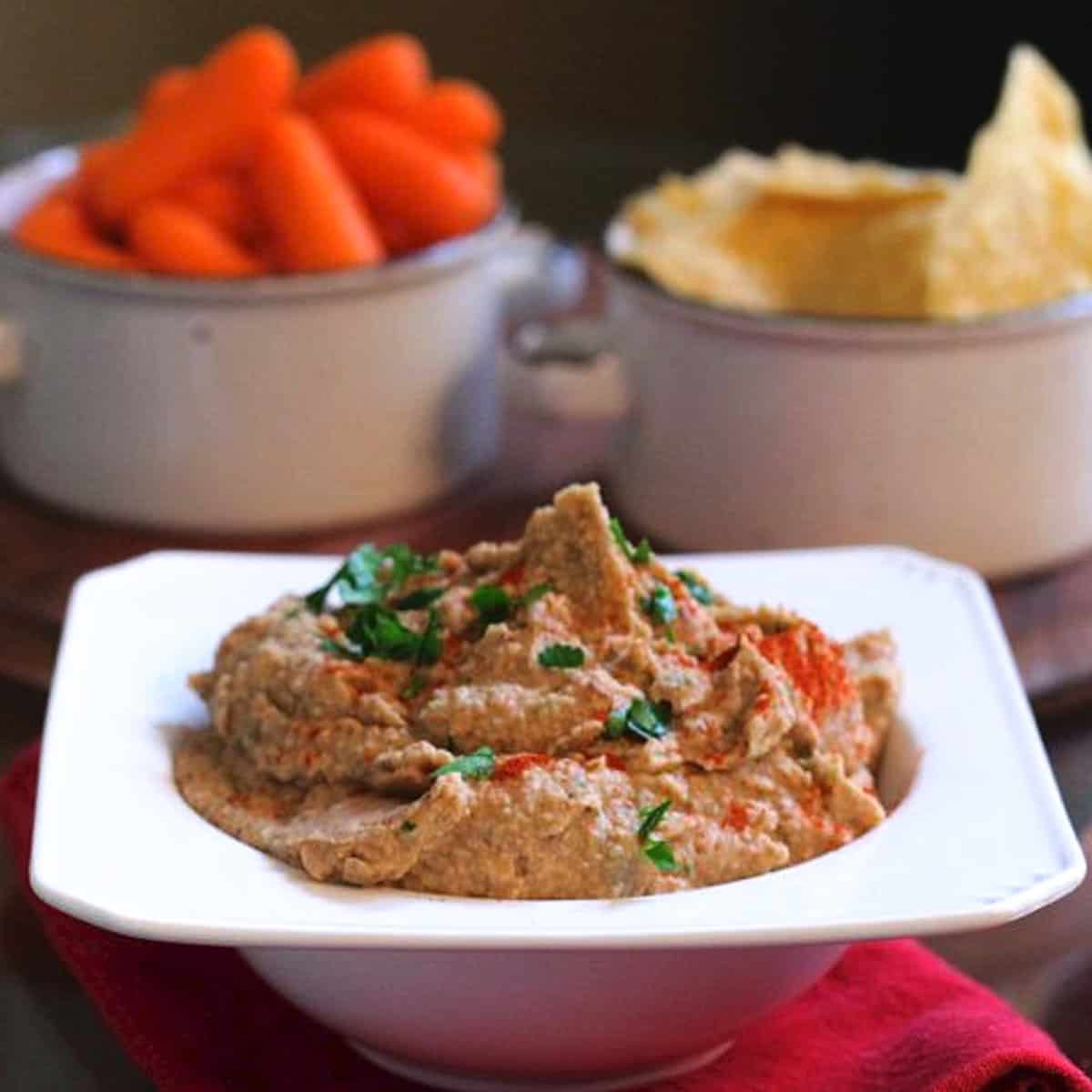 a bowl of black eyed pea hummus next to a red napkin and a bowl of carrots.