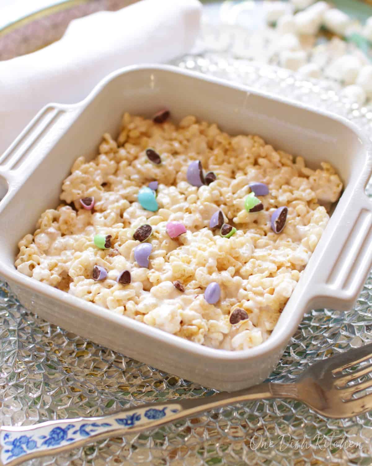 A giant rice krispies treat topped with pastel colored chocolate candies in a small baking dish next to a fork.