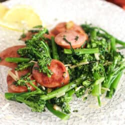 sauteed broccolini with sausage on a plate next to a slice of lemon.