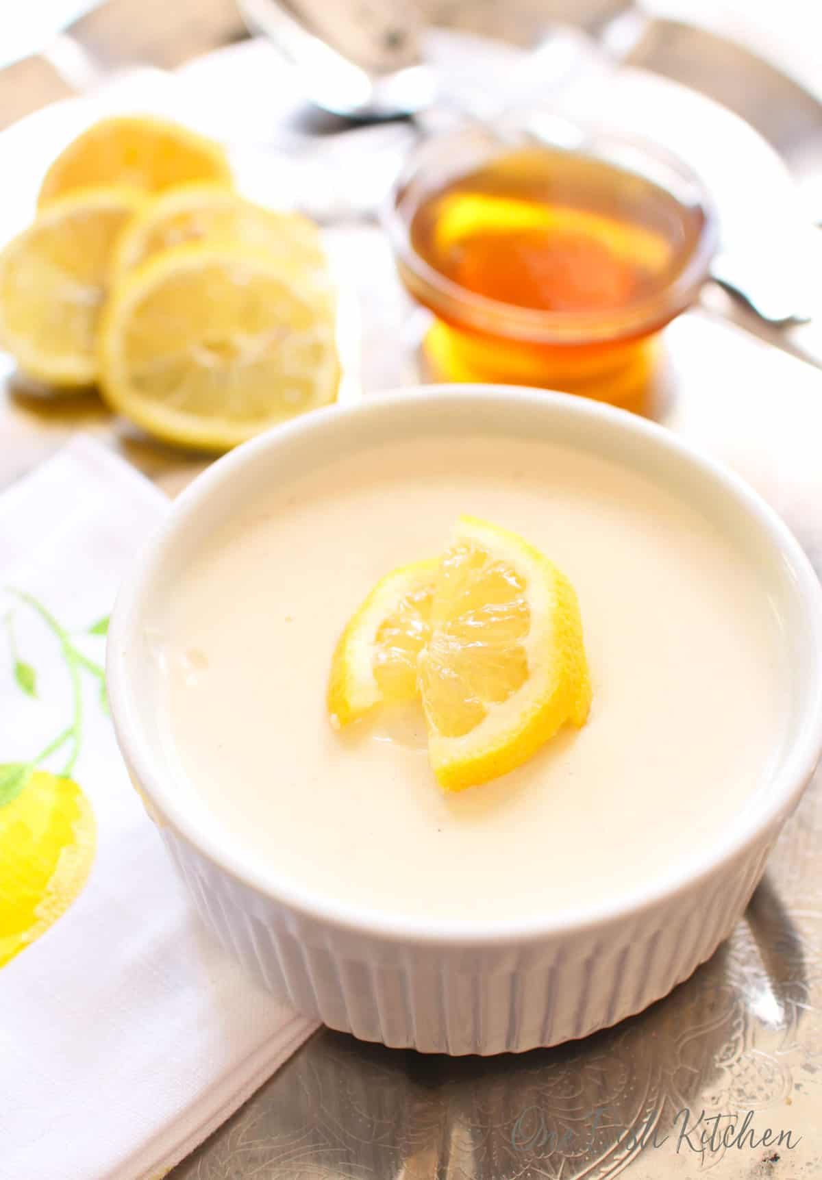 a small lemon posset next to a jar of honey and lemon slices on a silver tray.