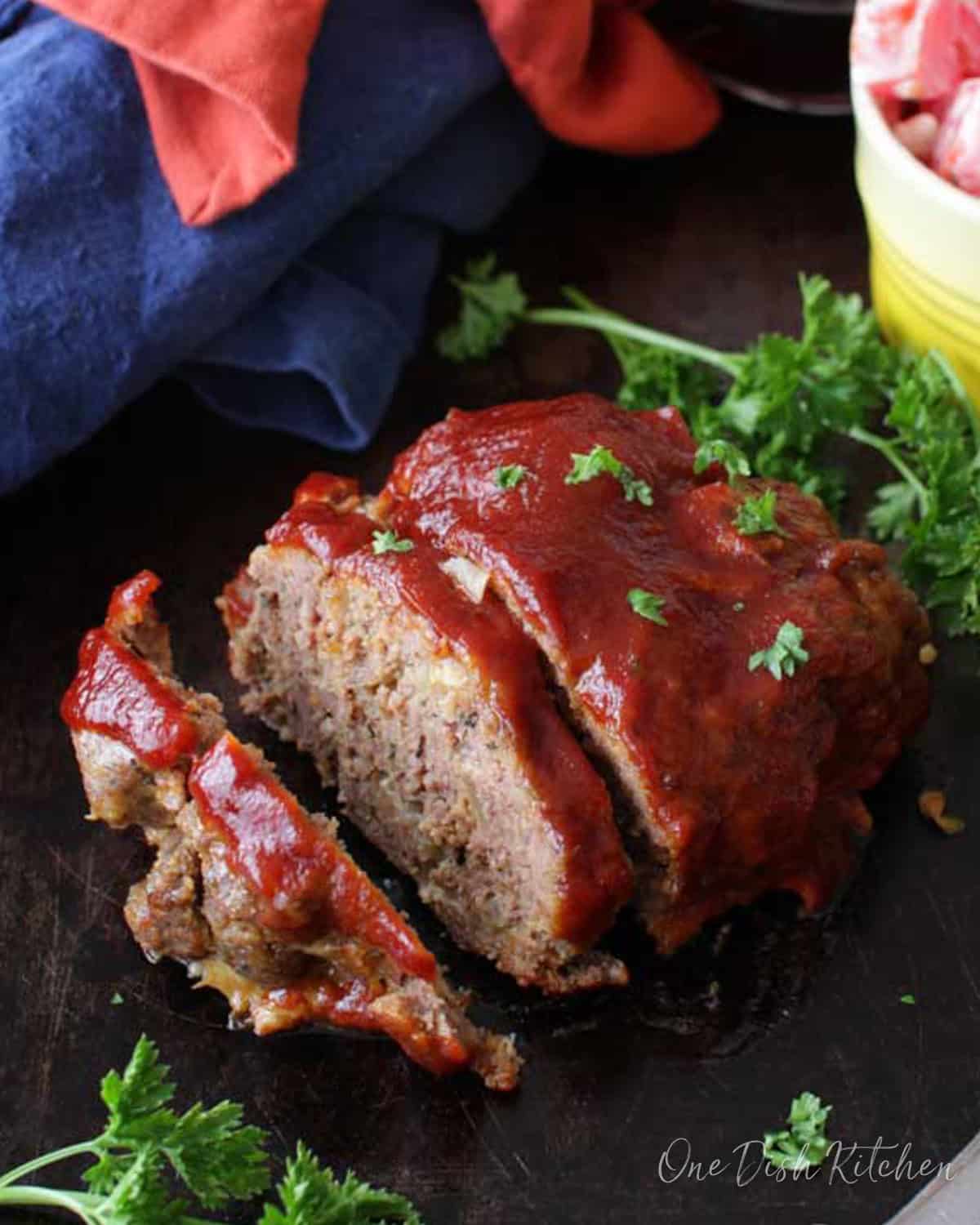 Meatloaf sliced on a dark wooden tray next to a small bowl of tomato slices with ranch dressing.