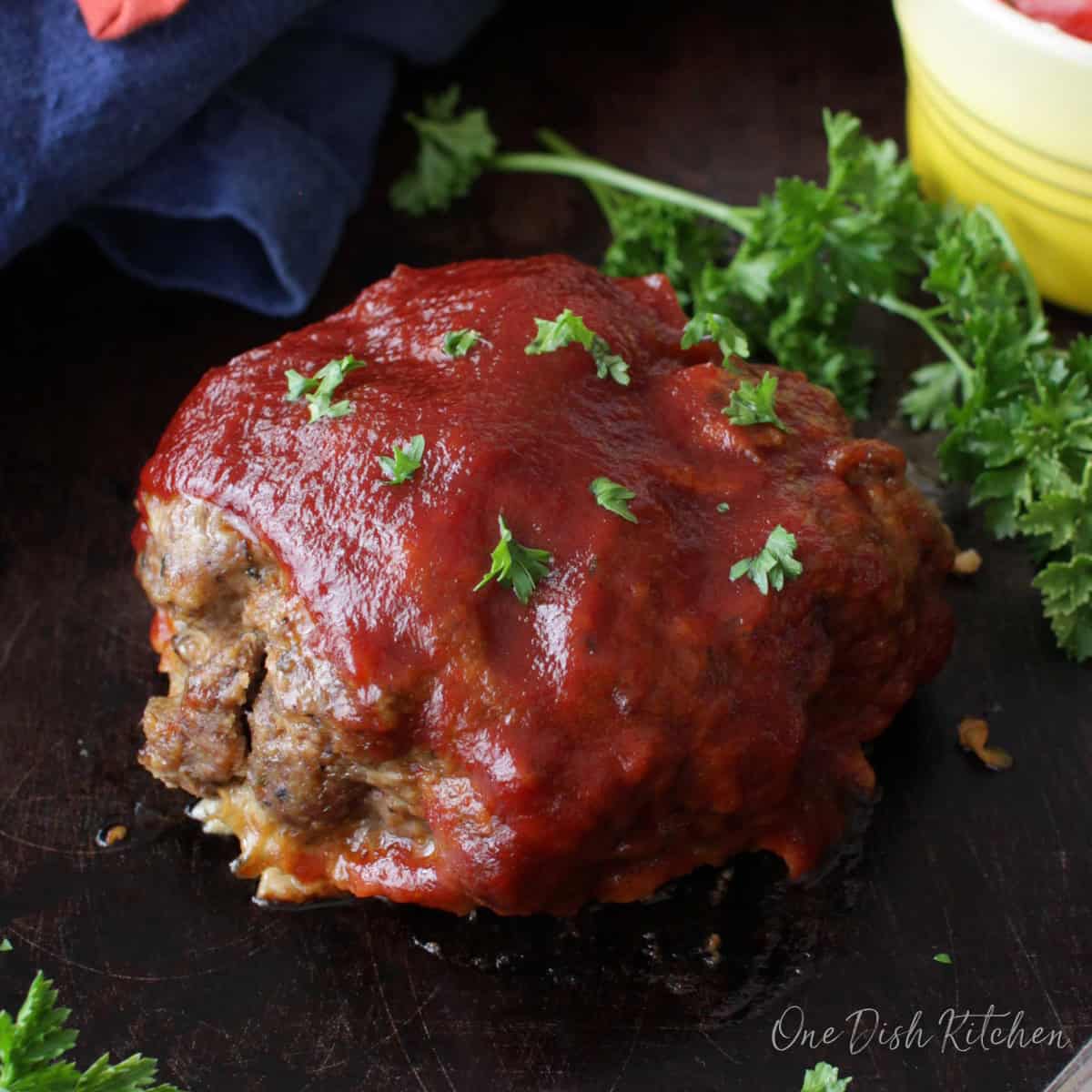 A single serving of meatloaf on a dark wooden tray with a glass of red wine and a small bowl of tomato slices with ranch dressing.
