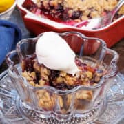 a small bowl of blueberry crumble topped with whipped cream next to a larger dish of the crumble.