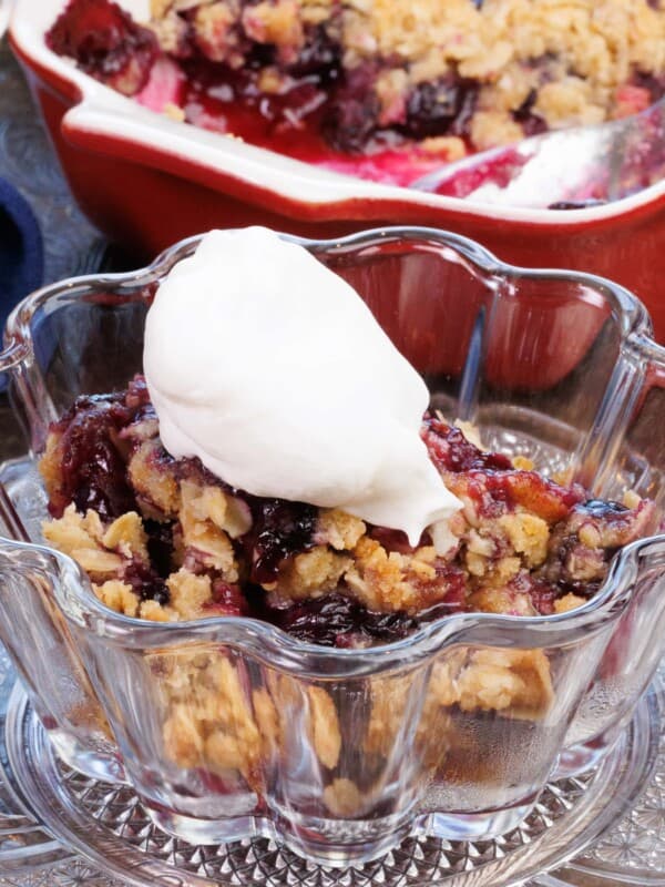 a small bowl of blueberry crumble topped with whipped cream next to a larger dish of the crumble.