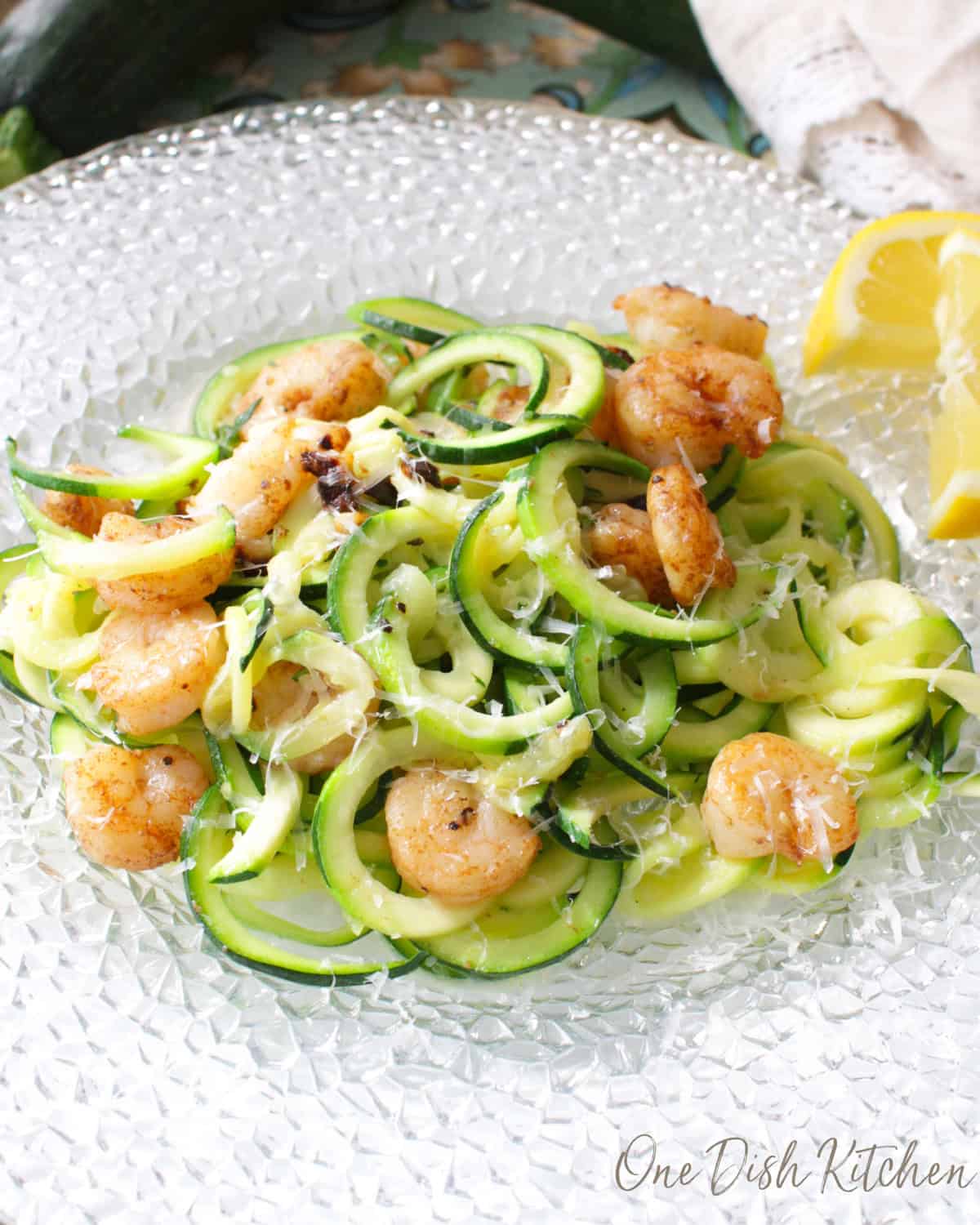 Zucchini noodles topped with grilled shrimp and parmesan cheese on a plate with lemon slices.