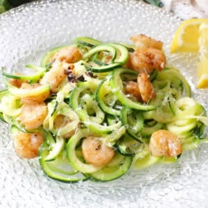 Zucchini noodles topped with grilled shrimp and parmesan cheese on a plate with lemon slices.