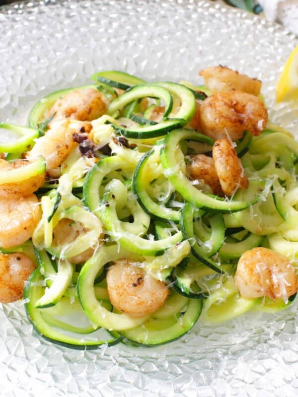 Zucchini noodles topped with grilled shrimp and parmesan cheese on a plate with lemon slices.