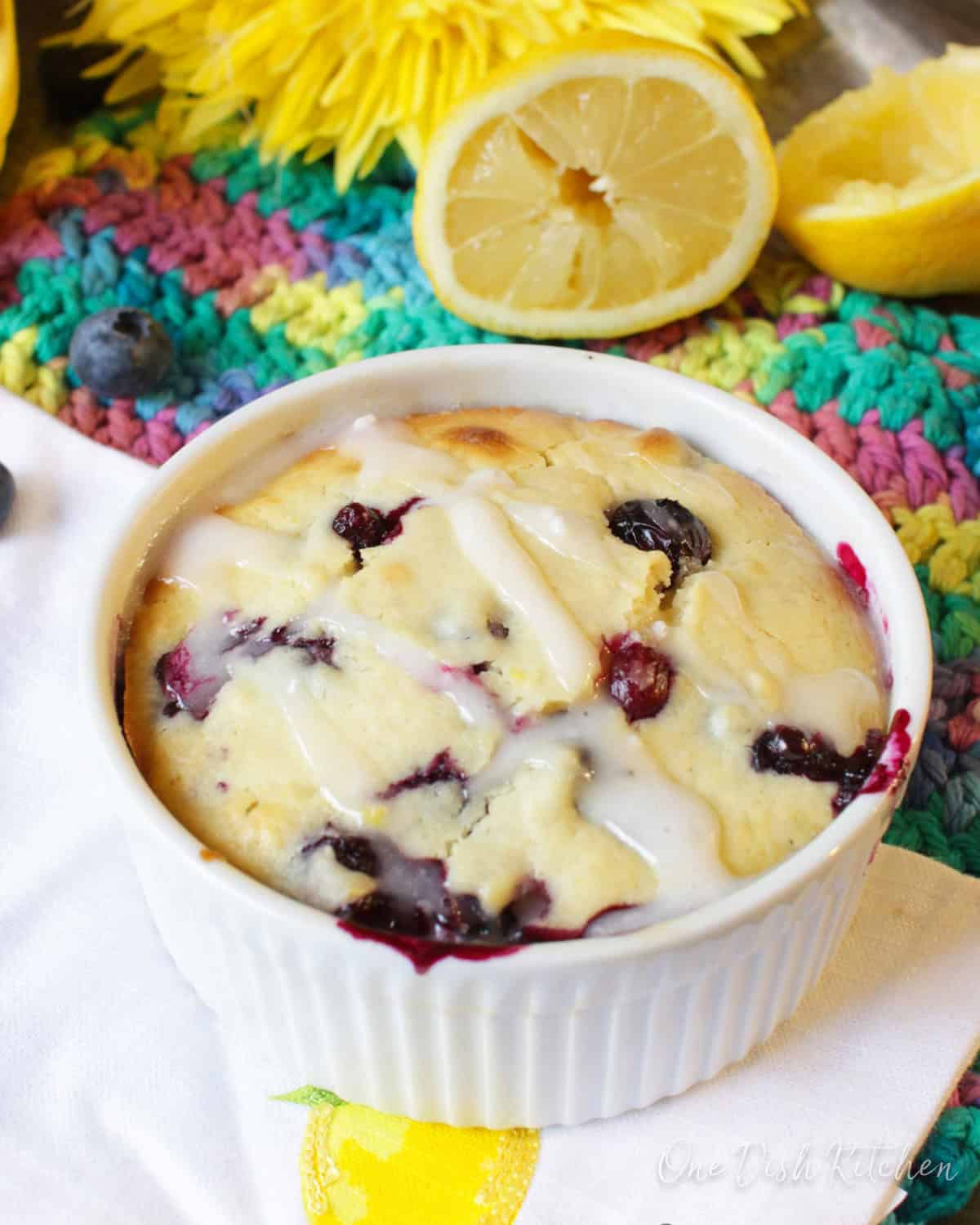 A lemon blueberry muffin in a ramekin topped with sugar glaze on a metal tray surrounded by blueberries, two lemon halves, and a big yellow flower.