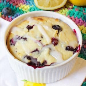 A lemon blueberry muffin in a ramekin topped with sugar glaze on a metal tray surrounded by blueberries, two lemon halves, and a big yellow flower.
