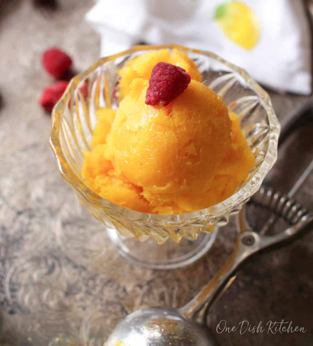 Mango sorbet topped with a raspberry in a dessert glass next to an ice cream scoop on a metal tray with scattered raspberries.
