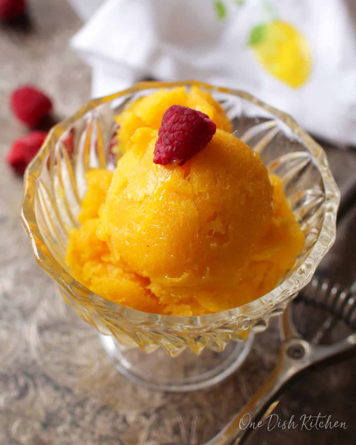 Mango sorbet topped with a raspberry in a dessert glass next to an ice cream scoop on a metal tray with scattered raspberries.