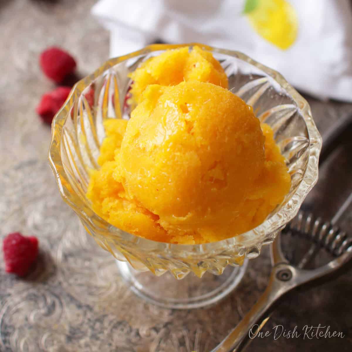 Mango sorbet in a dessert glass on a metal tray next to an ice cream scoop and scattered raspberries.