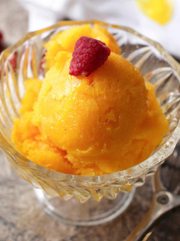 Mango sorbet topped with a raspberry in a dessert glass next to an ice cream scoop on a metal tray with scattered raspberries.