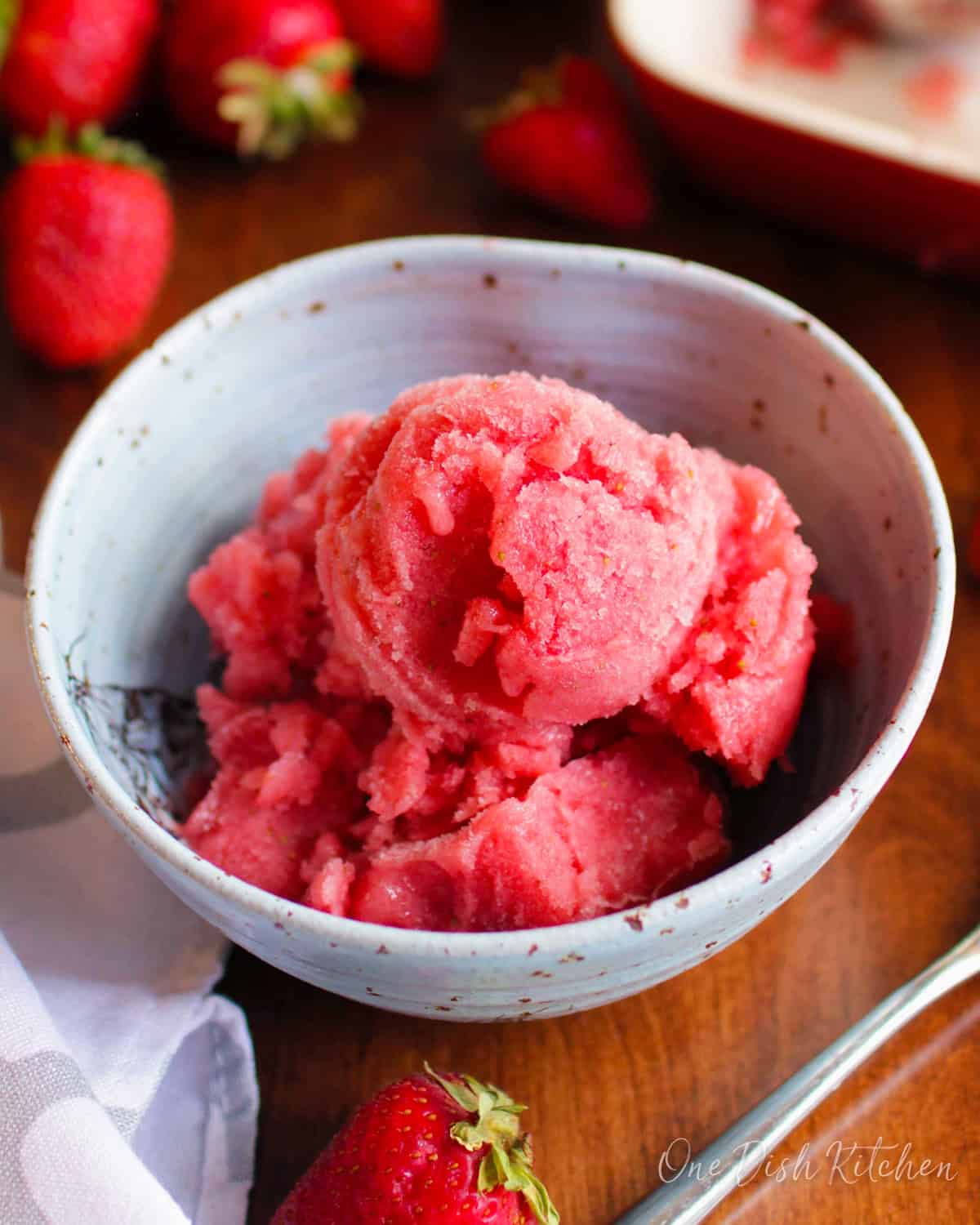 a blue bowl filled with watermelon sorbet surrounded by fresh strawberries on a wooden table.