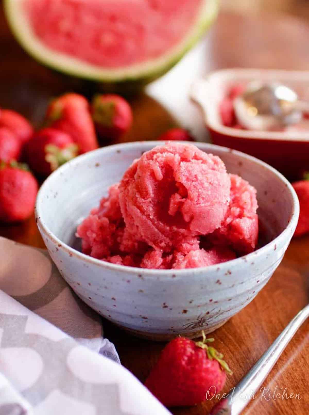 a blue bowl filled with watermelon sorbet surrounded by fresh strawberries on a wooden table.