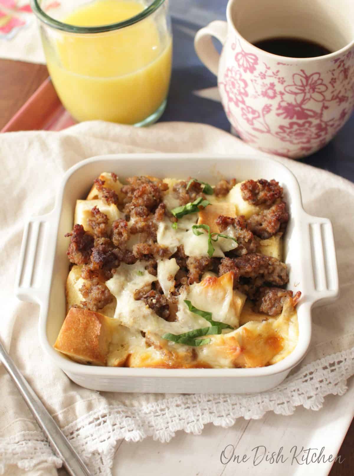 Breakfast casserole in a small baking dish next to a glass of orange juice and a coffee mug.