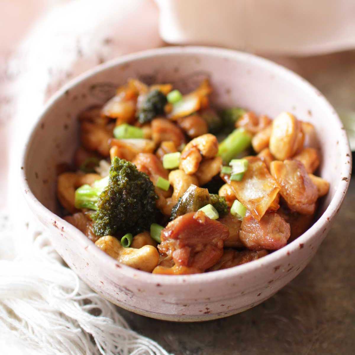 a pink bowl filled with cashew chicken and broccoli next to a pink lace napkin.