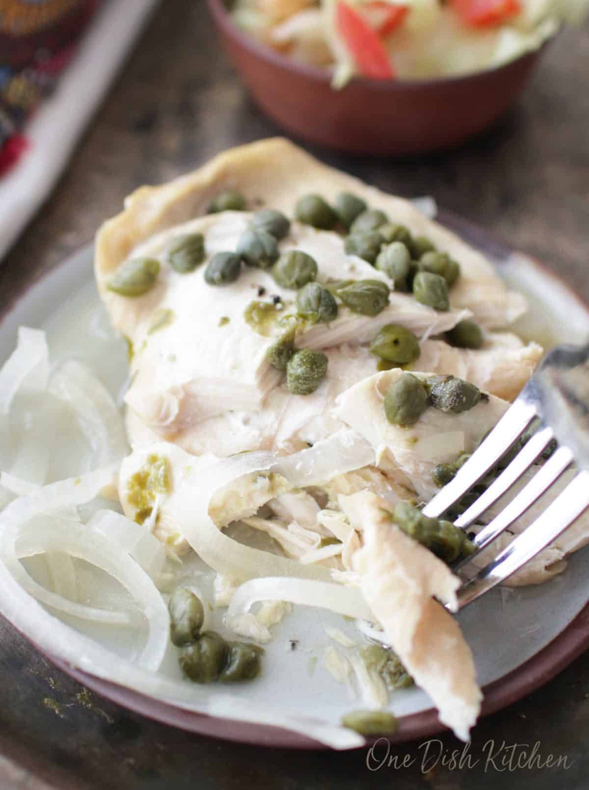 a piece of chicken topped with capers and a fork scooping some of the chicken off the plate.