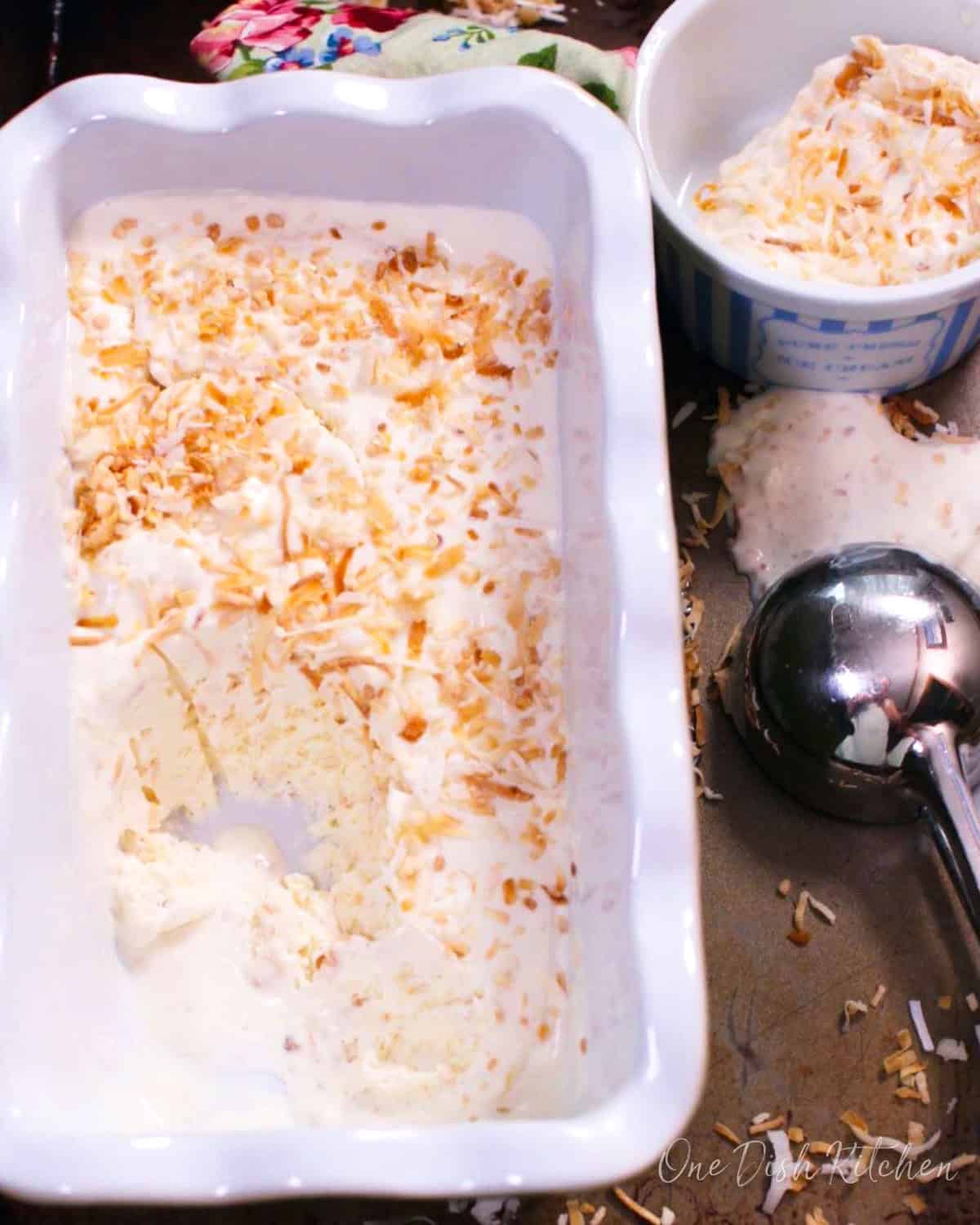 Toasted coconut ice cream in a loaf pan next to an ice cream scoop and a stack of two ice cream bowls.