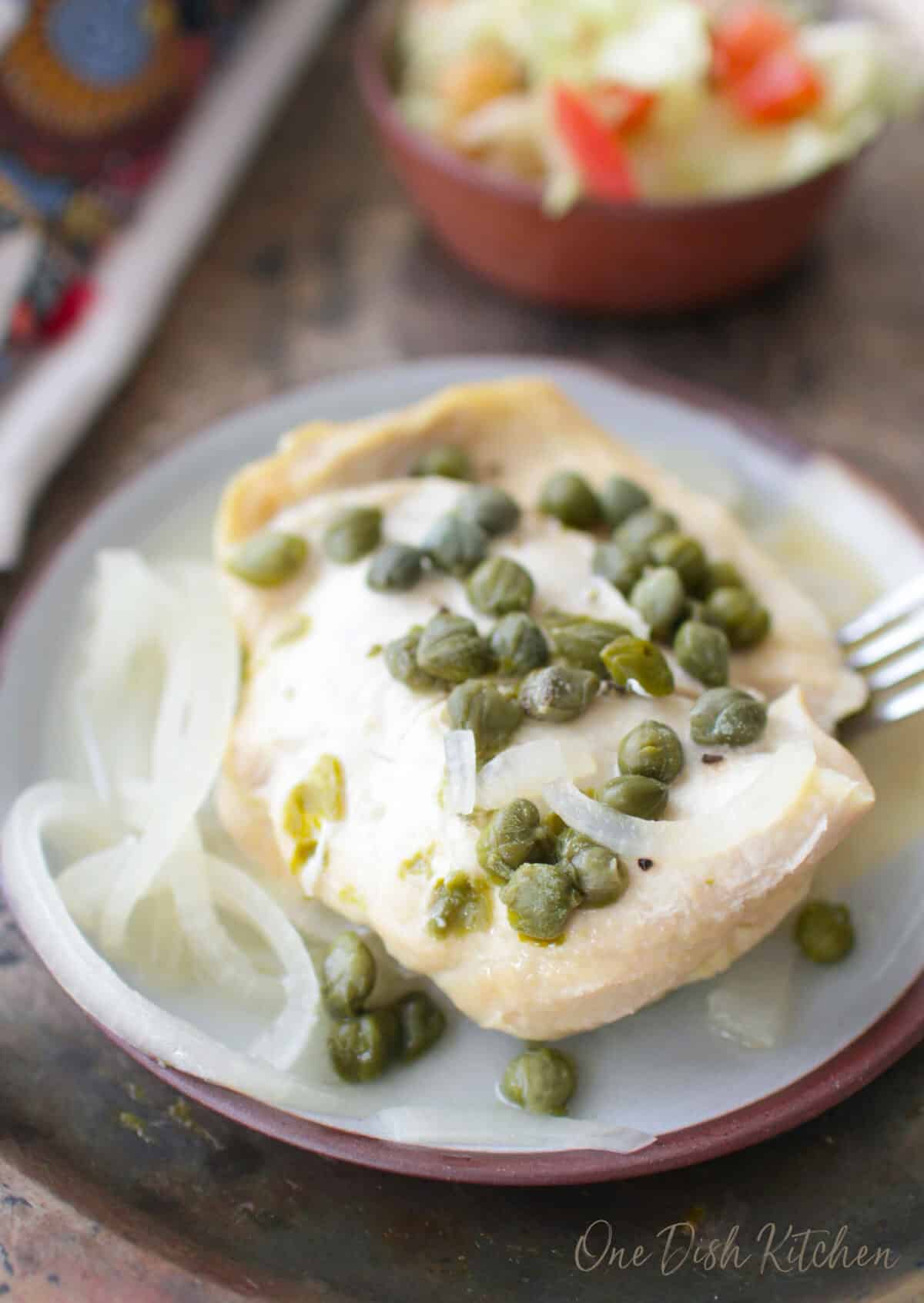 a chicken breast topped with slices of white onions and capers on a blue plate.