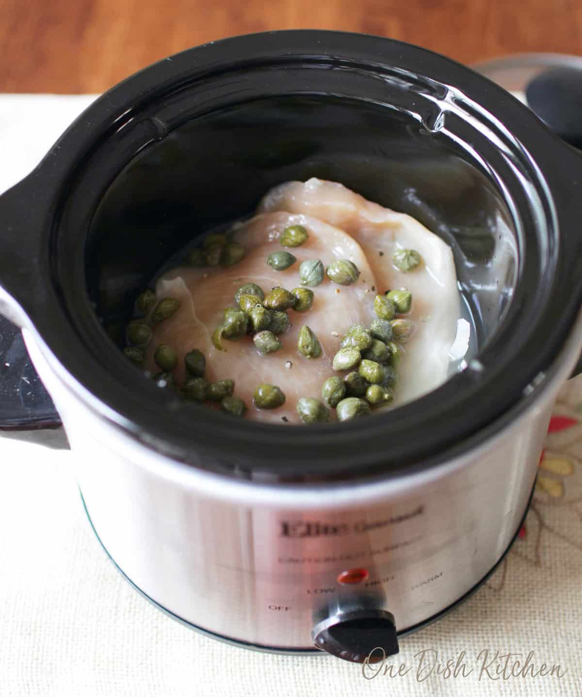 a chicken breast in a small slow cooker topped with sliced onions, broth, and capers.