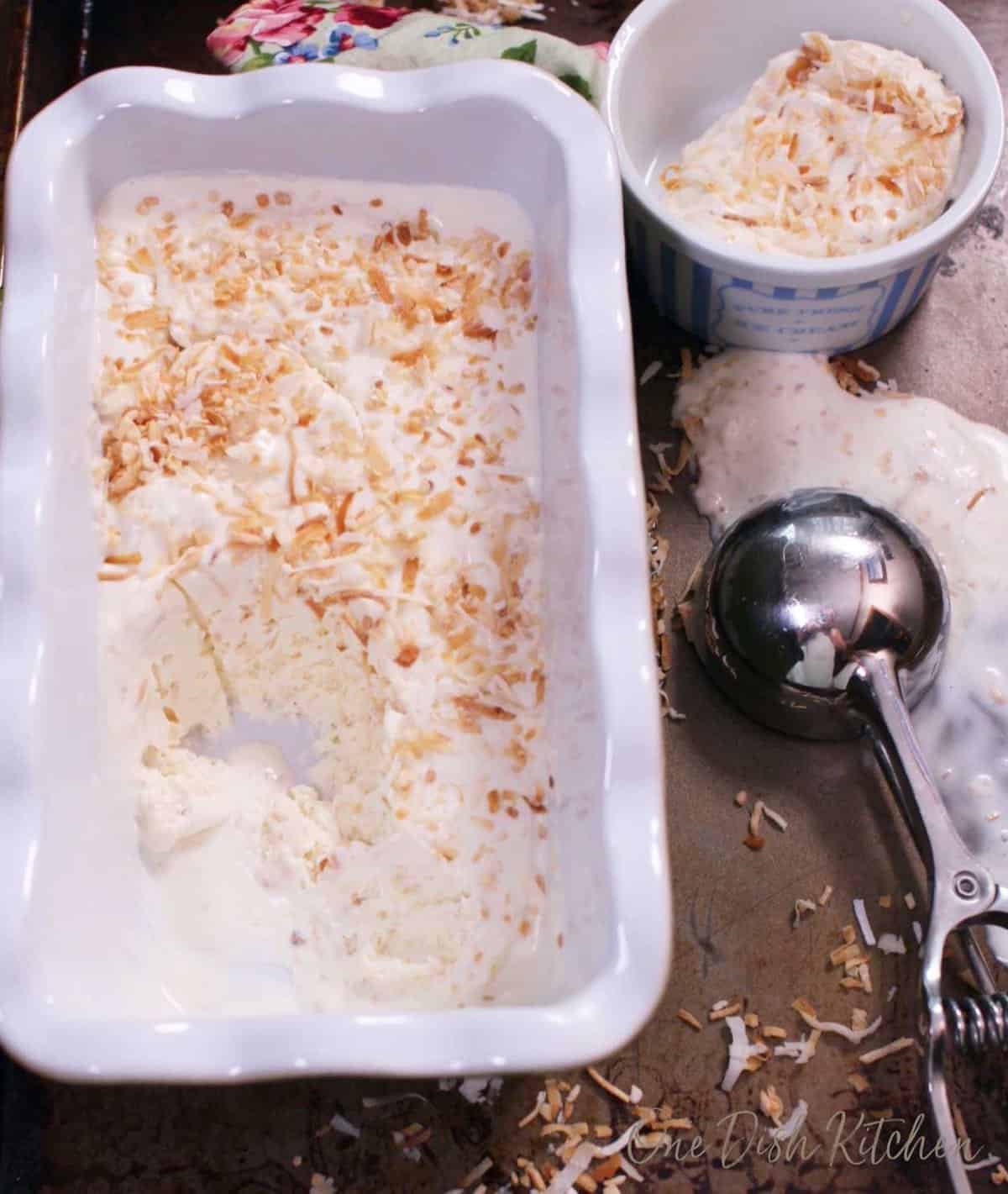 Toasted coconut ice cream in a loaf pan next to an ice cream scoop and a stack of two ice cream bowls.