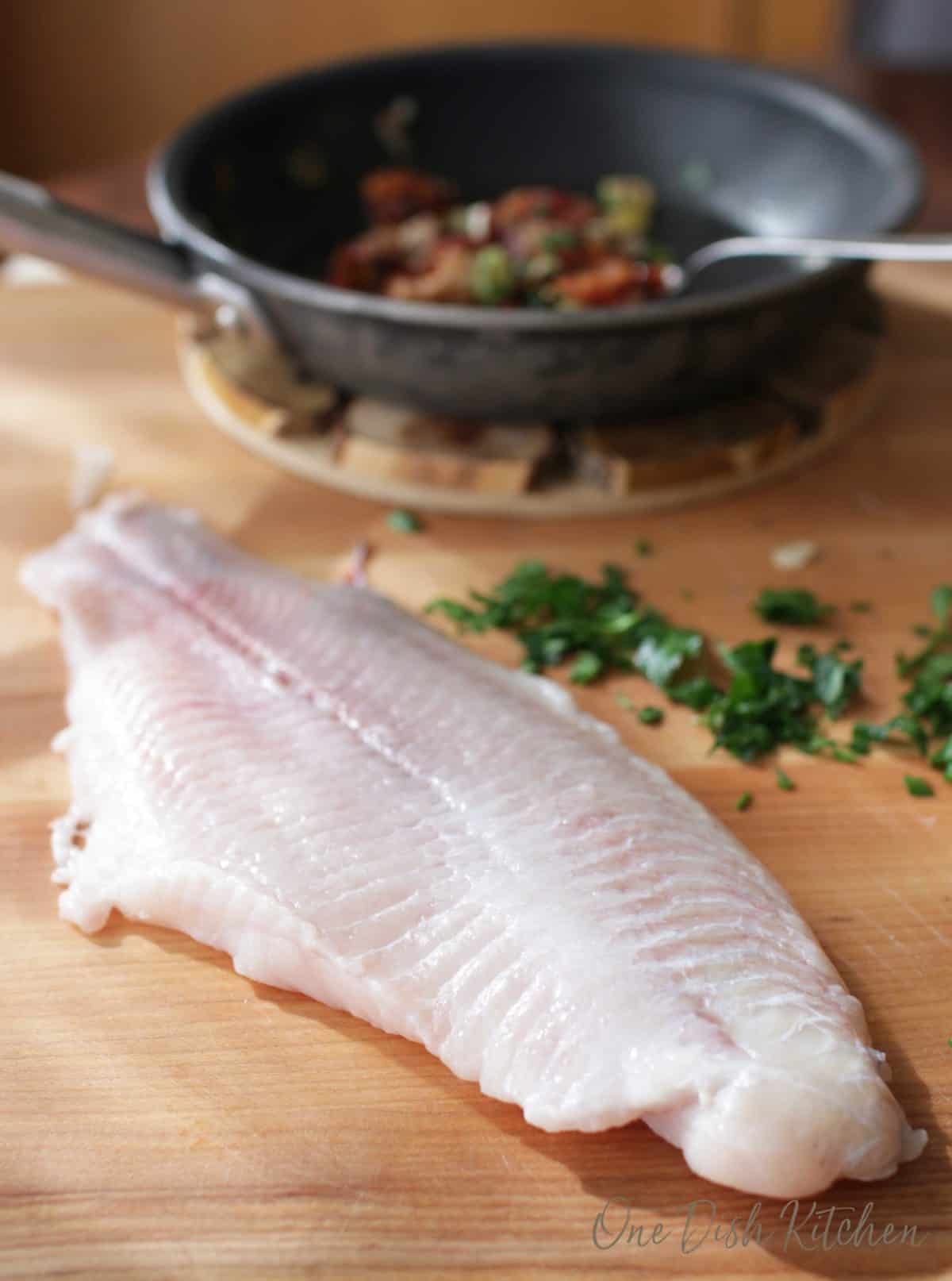 a catfish fillet on a brown cutting board with a skillet in the background filled with chopped bacon and vegetables.
