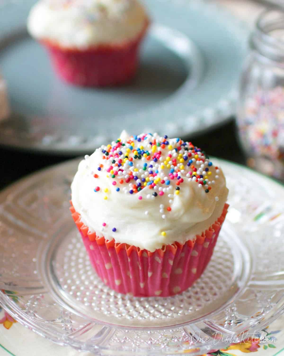 two vanilla cupcakes on plates topped with vanilla frosting and next to a jar of sprinkles.