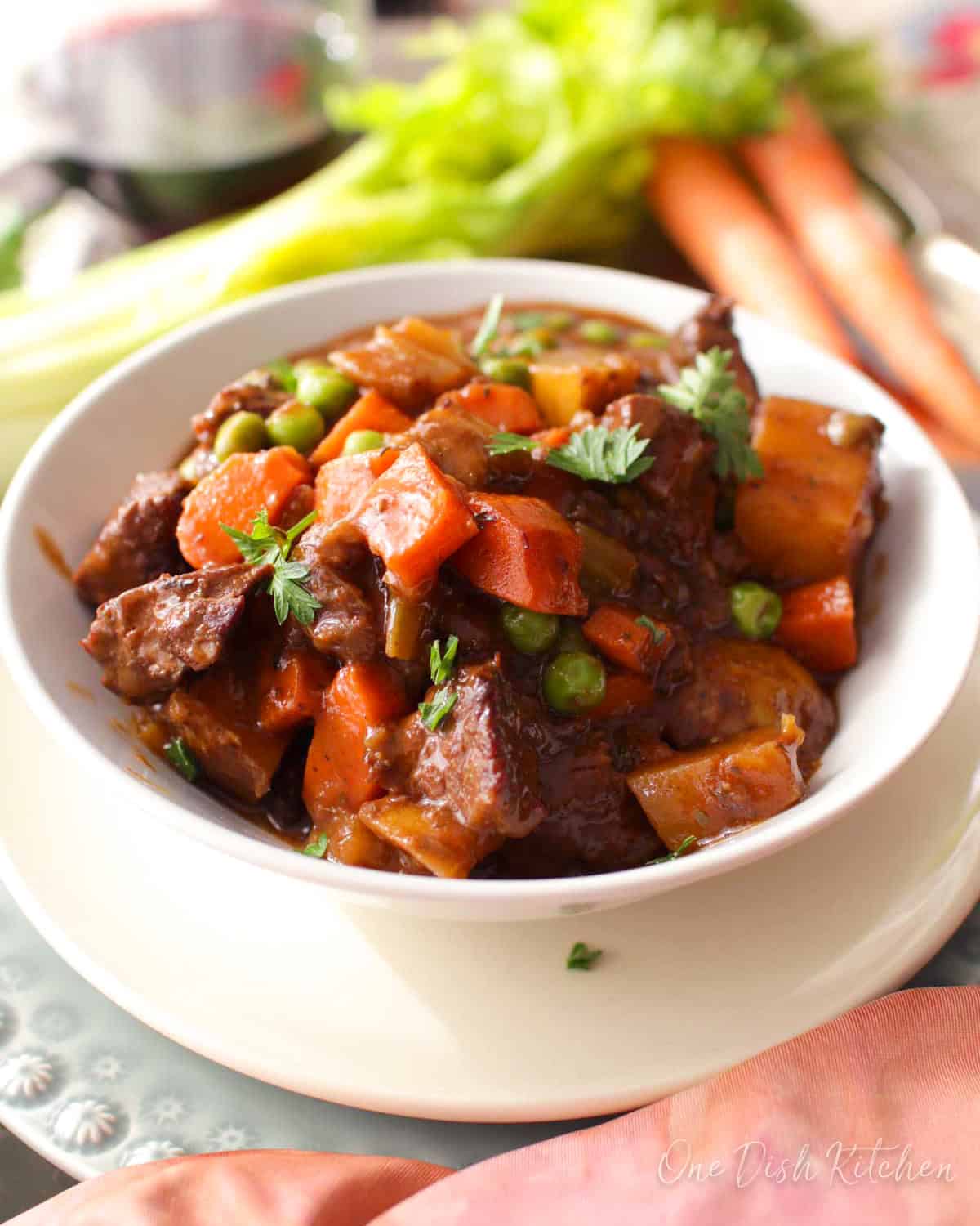 A single serving of beef stew in a white bowl next to fresh carrots.
