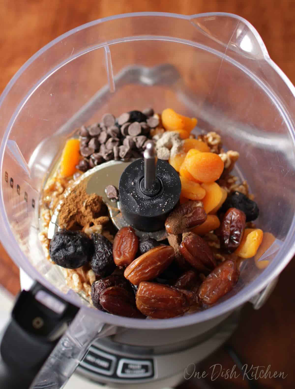 dates, dried plums, dried apricots, chocolate chips and walnuts in the base of a food processor on a brown table.