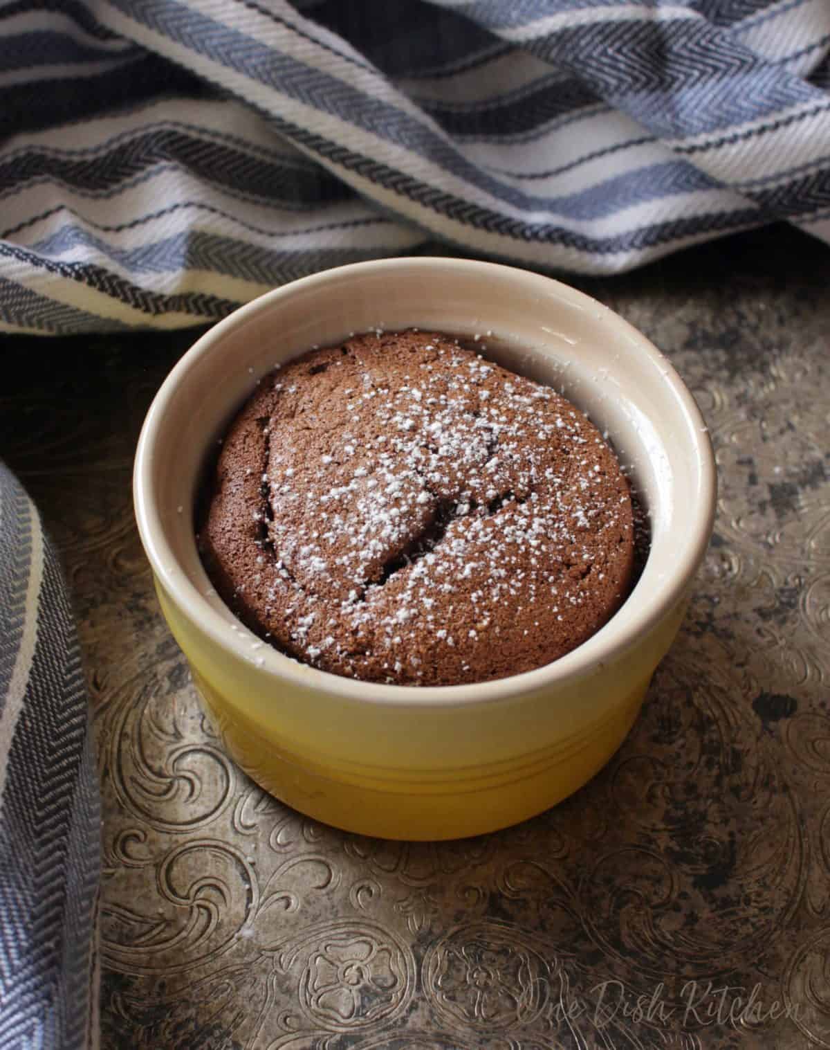 a mini chocolate cake in a yellow baking dish on a silver tray next to a blue and white kitchen towel.