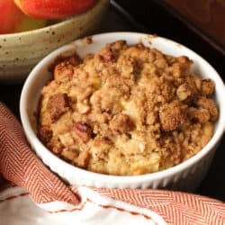a bakery style apple muffin topped with a pecan streusel topping next to a bowl of apples and an orange napkin