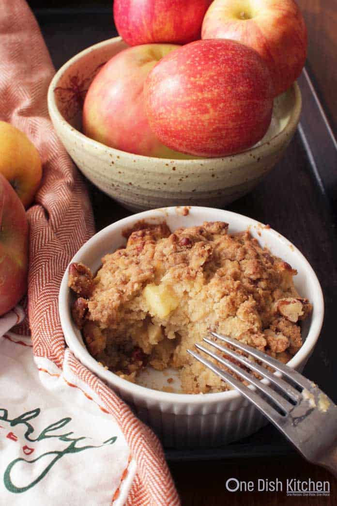 A forkful missing from an apple muffin in a ramekin topped with cinnamon streusel with a fork resting on the side all next to a bowl of apples