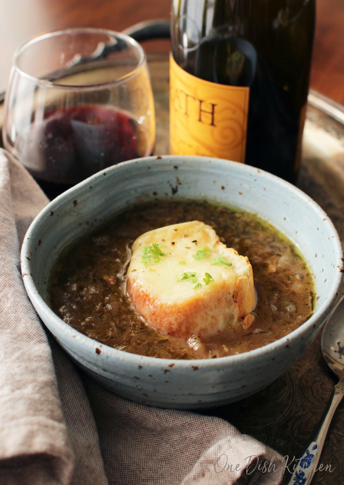 A bowl of french onion soup on a metal tray with a glass and bottle of red wine and a spoon.
