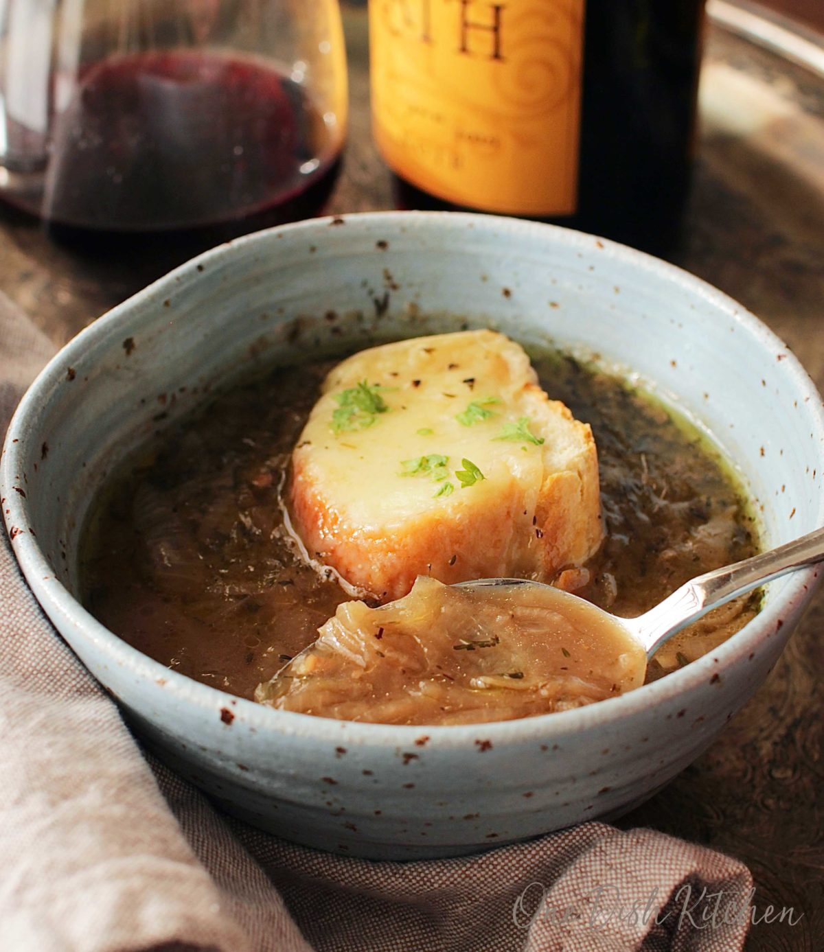 A spoonful of french onion soup from a bowl on a metal tray with a glass and bottle of red wine.