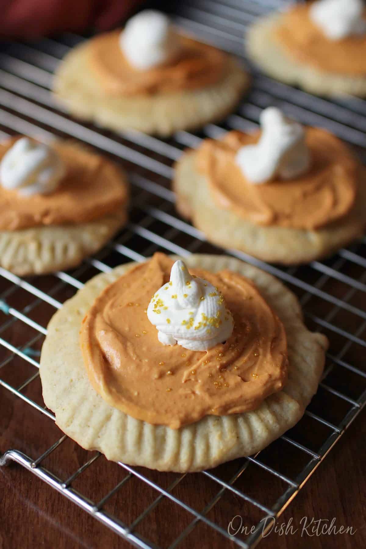 Tray of pumpkin cookies topped with orange frosting and a dollop of white icing on cooling on a rack.