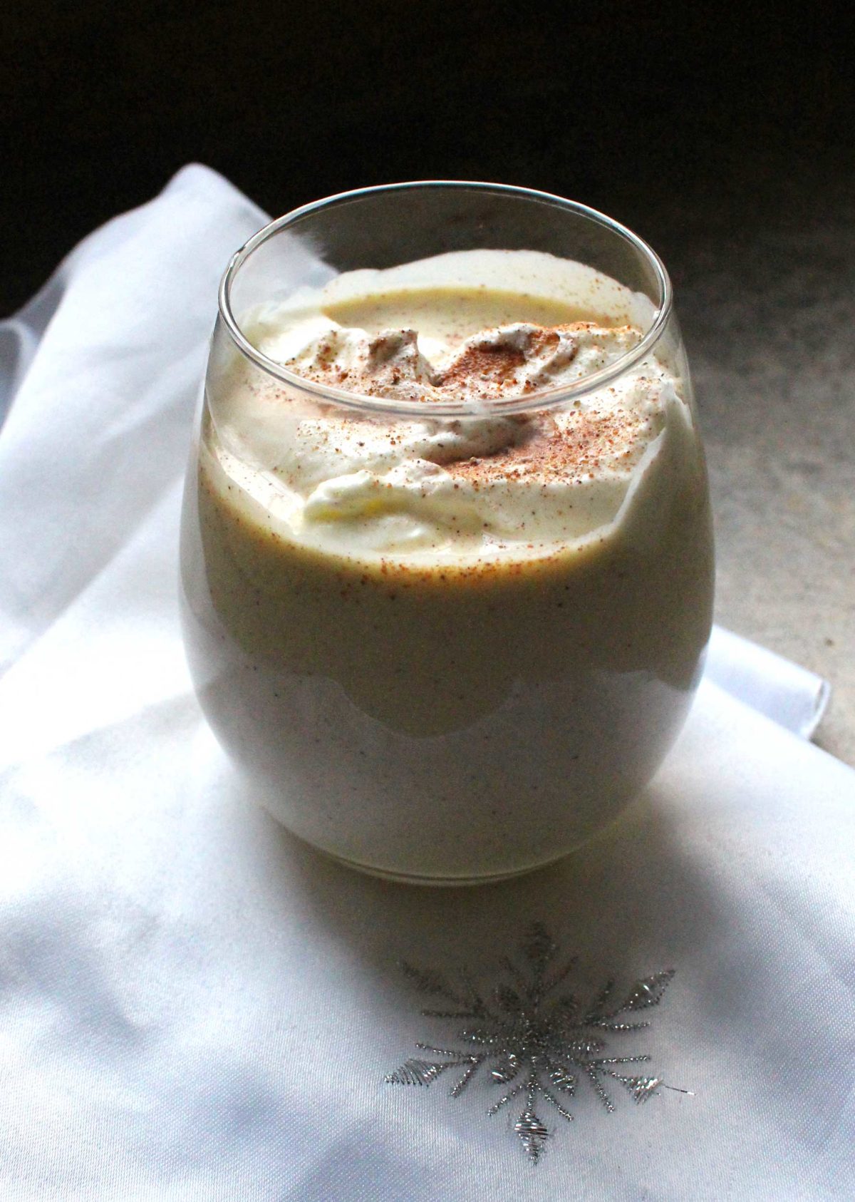 a glass of eggnog topped with whipped cream and nutmeg on a white napkin.
