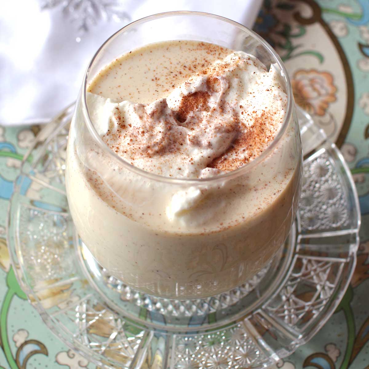 a glass of eggnog topped with whipped cream and nutmeg on a white napkin.