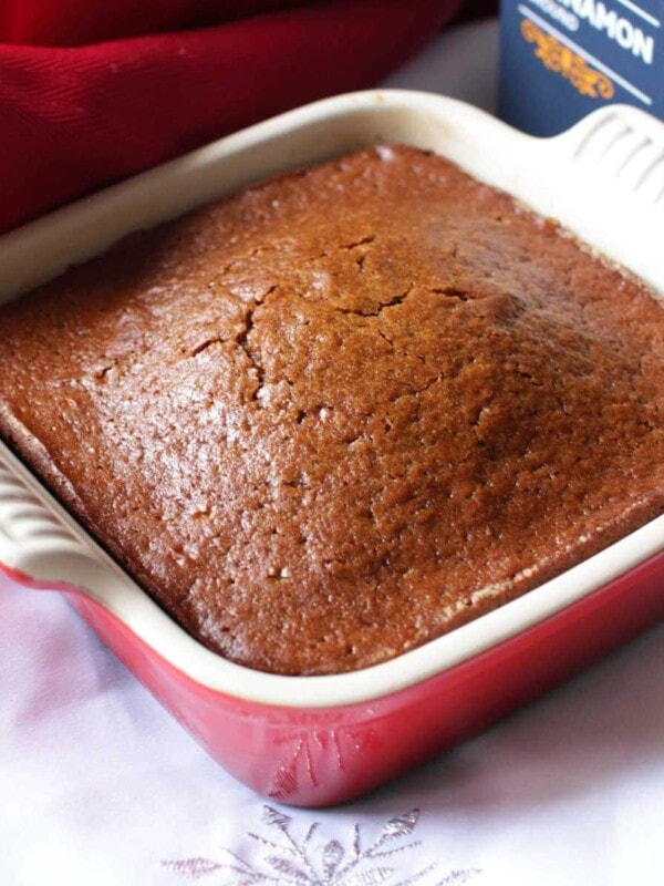 a gingerbread cake in a square red baking dish on a white napkin with a container of cinnamon in the background