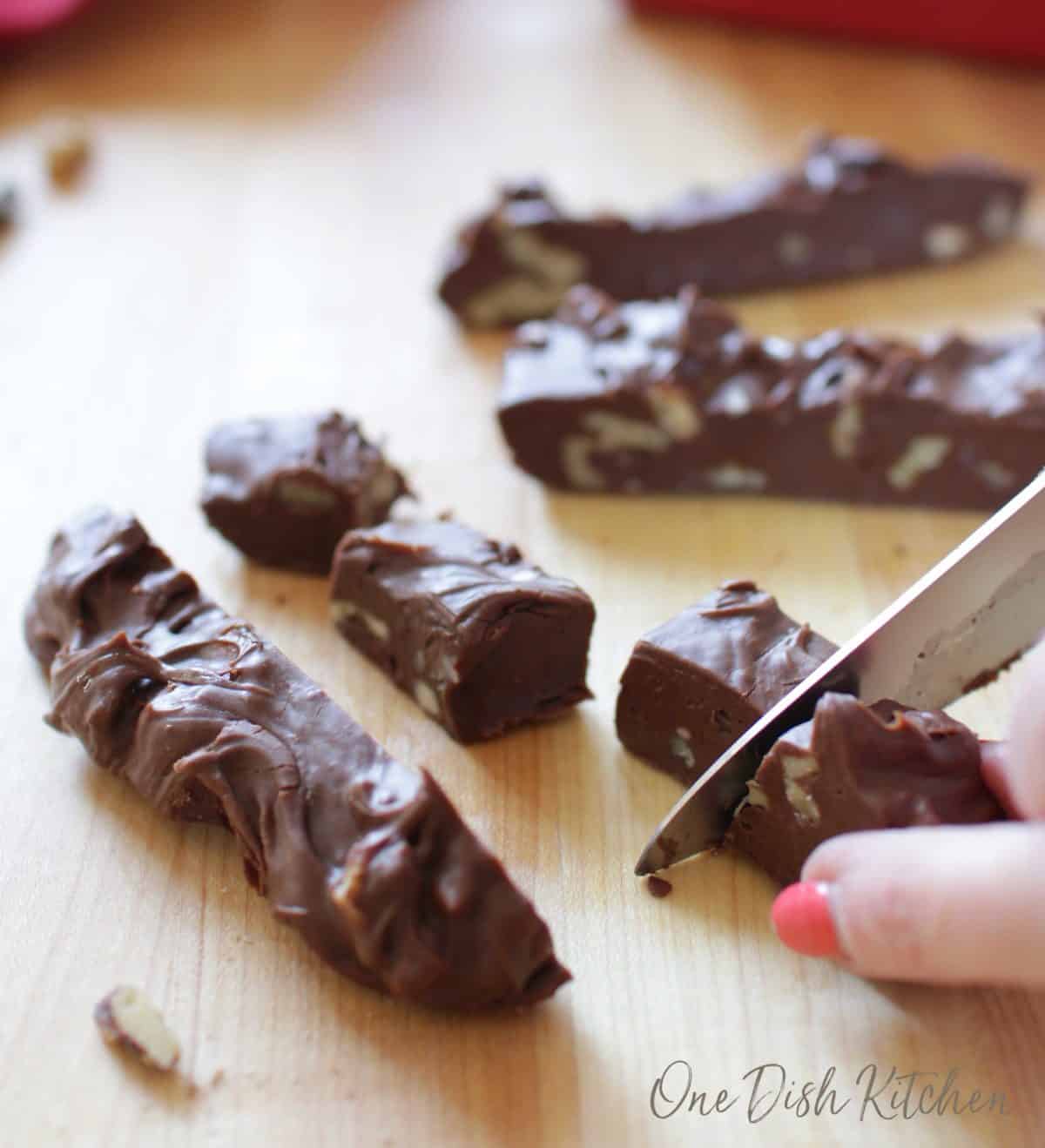 slicing 4 strips of fudge into squares on a brown cutting board.