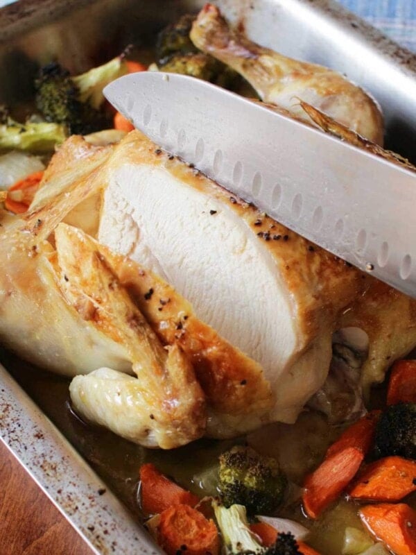 a whole roasted chicken in a large roasting pan being sliced with a large knife