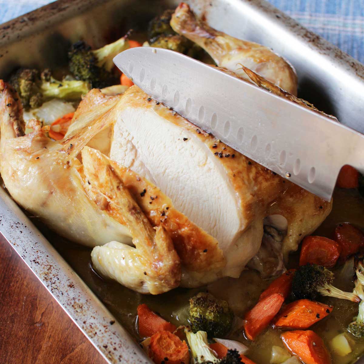 a whole roasted chicken in a large roasting pan being sliced with a large knife