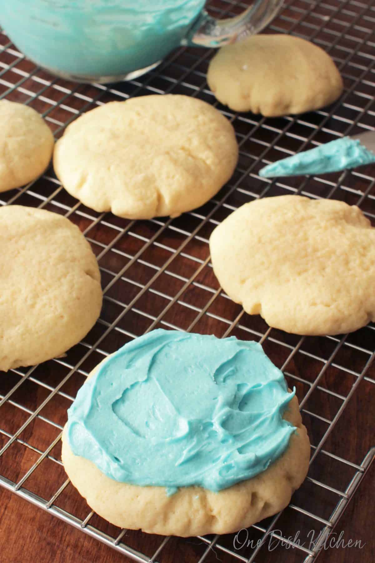 Six Sugar Cookies on a cooling rack with one cookie topped with blue frosting next to a glass bowl of blue frosting 
